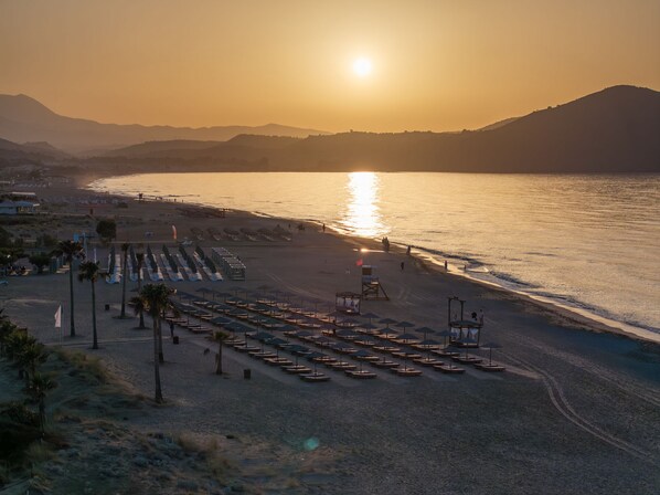 A beach with rows of sun loungers and umbrellas, a lifeguard tower, and a sunset over the water. - Delfina Tropic Beach (Apokoronas)