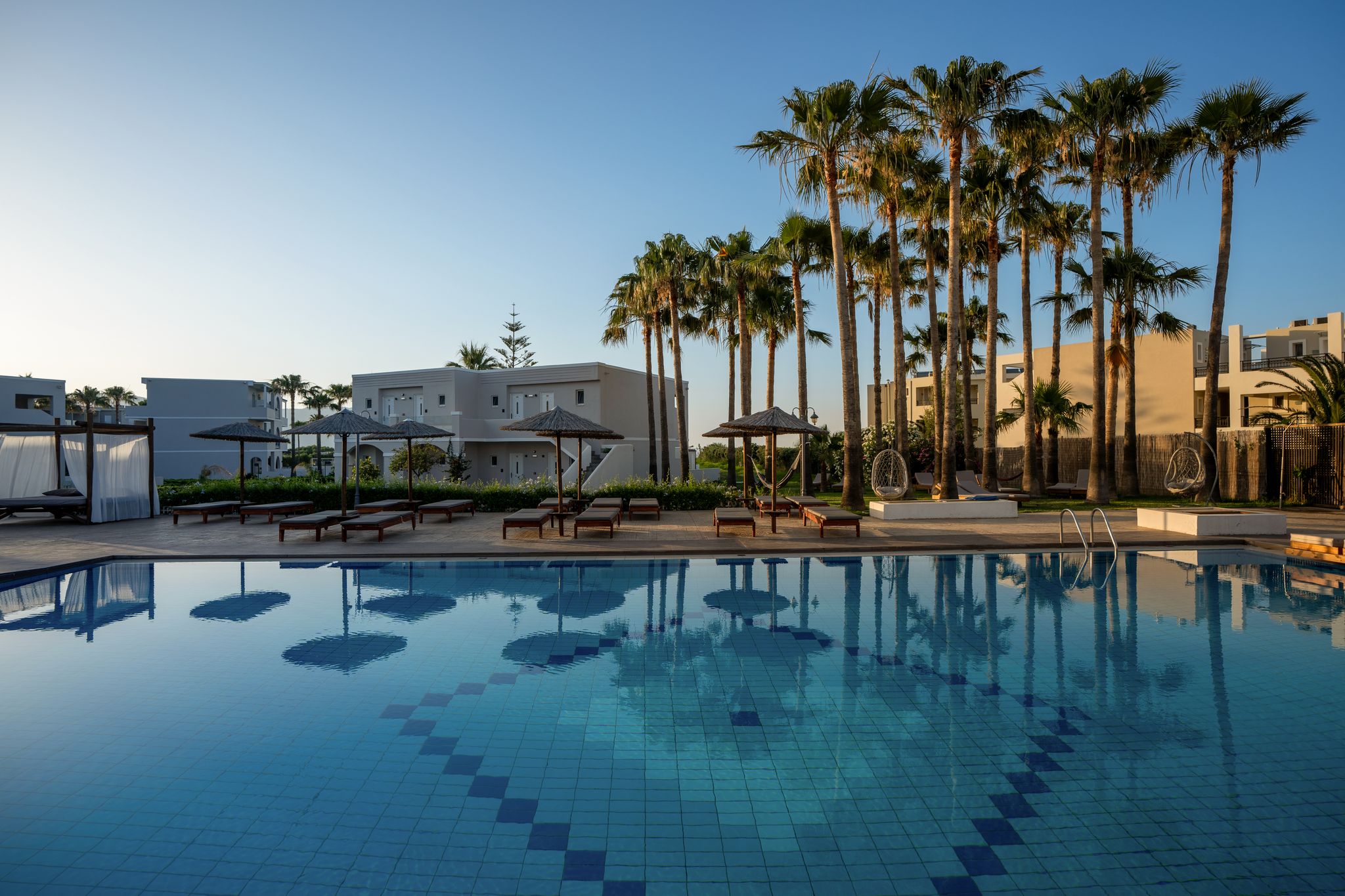 Une piscine entourée de palmiers et de bâtiments modernes, avec des chaises longues et des parasols.