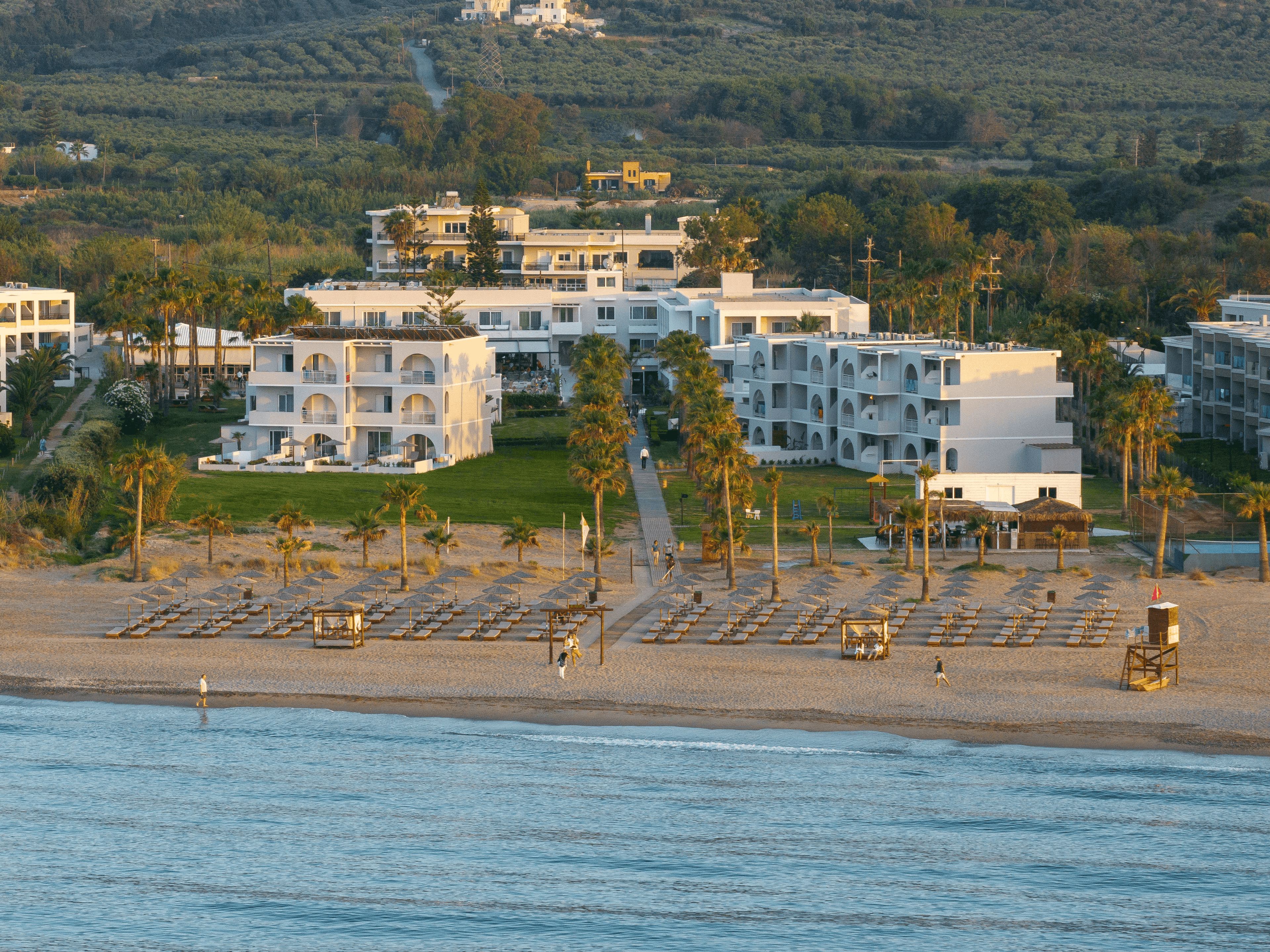Un hotel frente al mar con varias construcciones, una playa con tumbonas y vistas al océano.