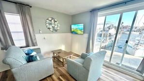 Een woonkamer met twee grijze fauteuils, een houten salontafel en een aan de muur bevestigde televisie.