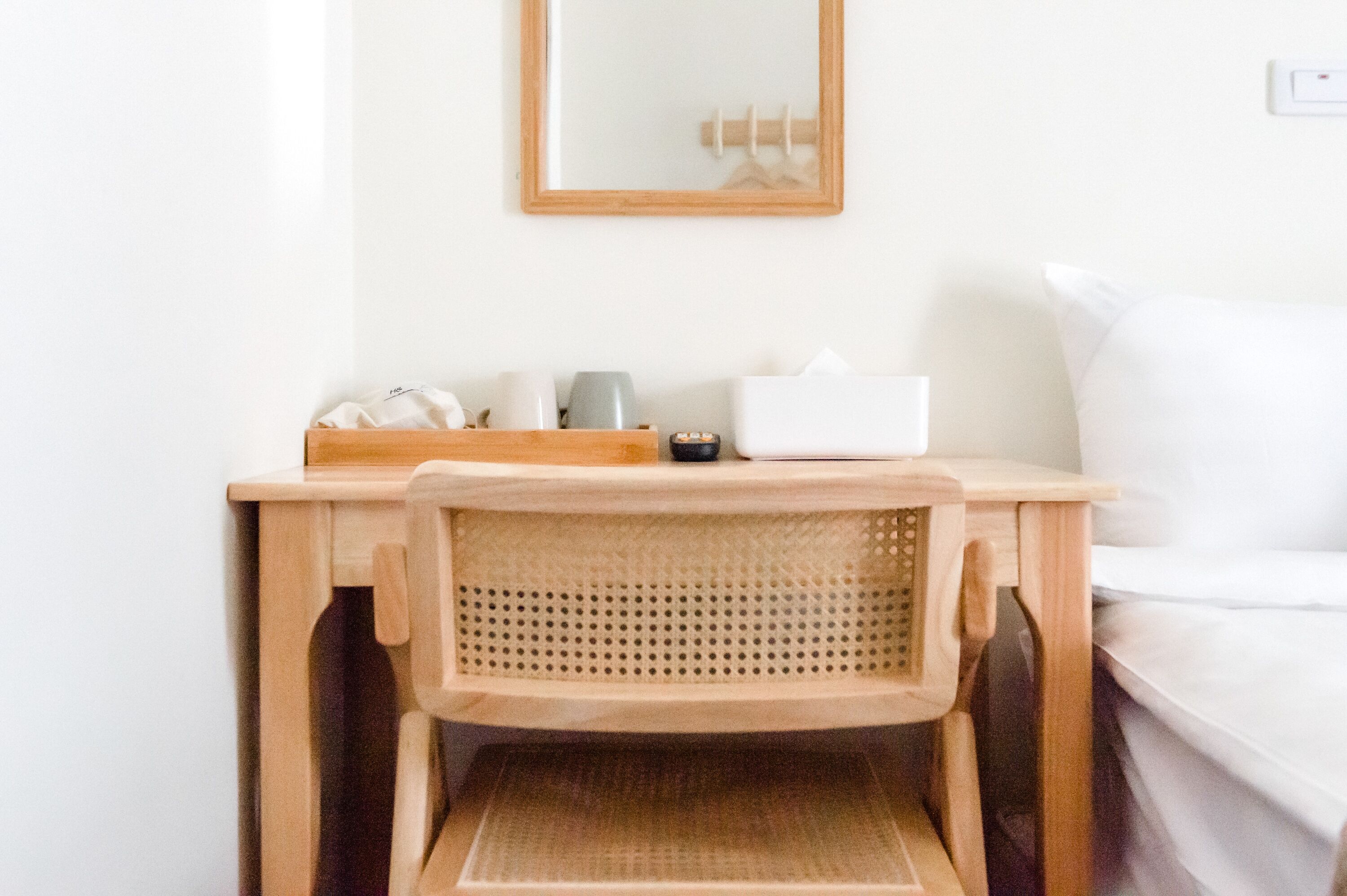 A wooden desk with a woven chair, a mirror, and a white bedside table with a white pillow.