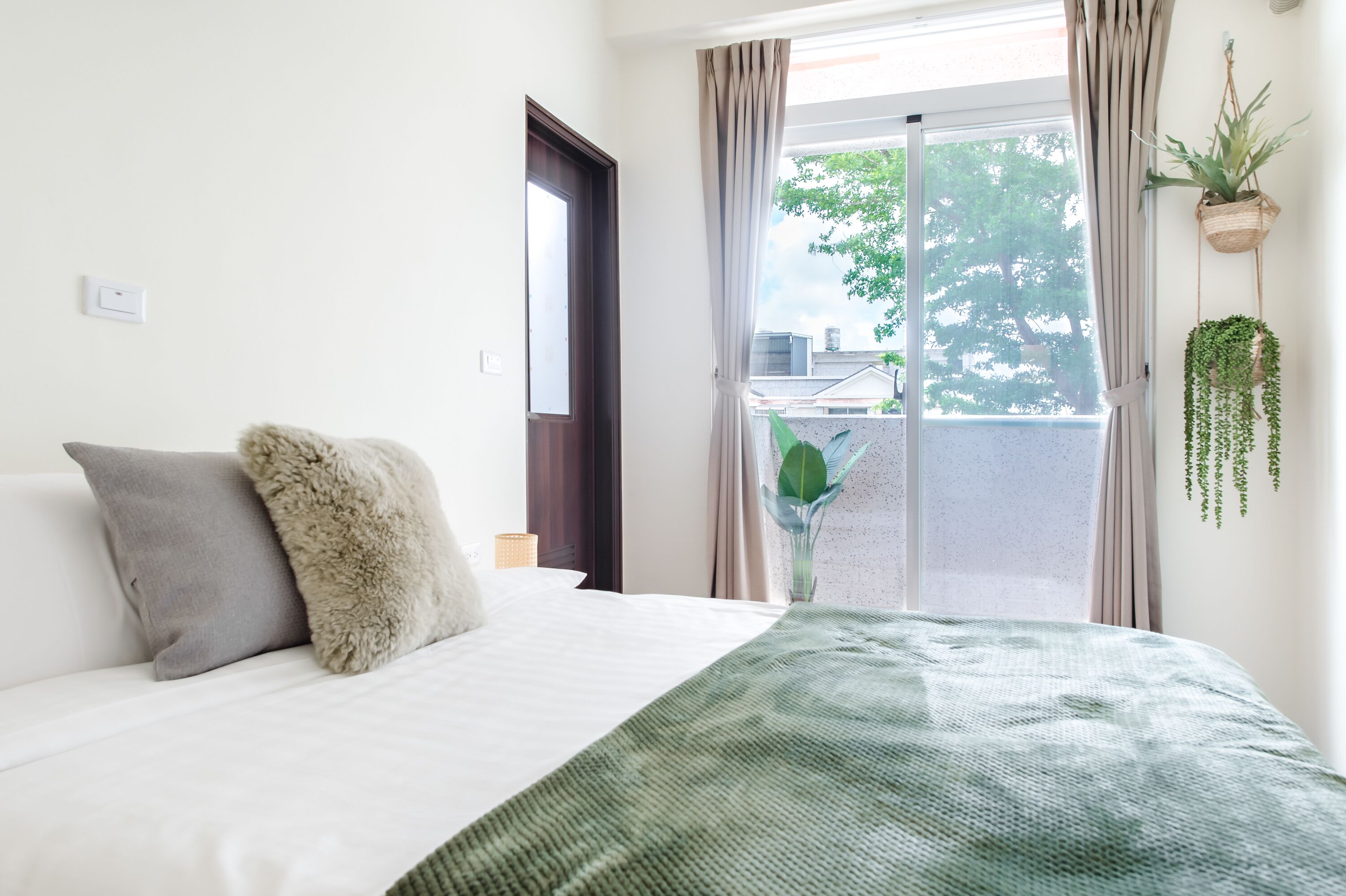 A neatly made bed with a green blanket, a fluffy pillow, and a grey pillow. There is a window with curtains, a potted plant, and a wall-mounted plant.