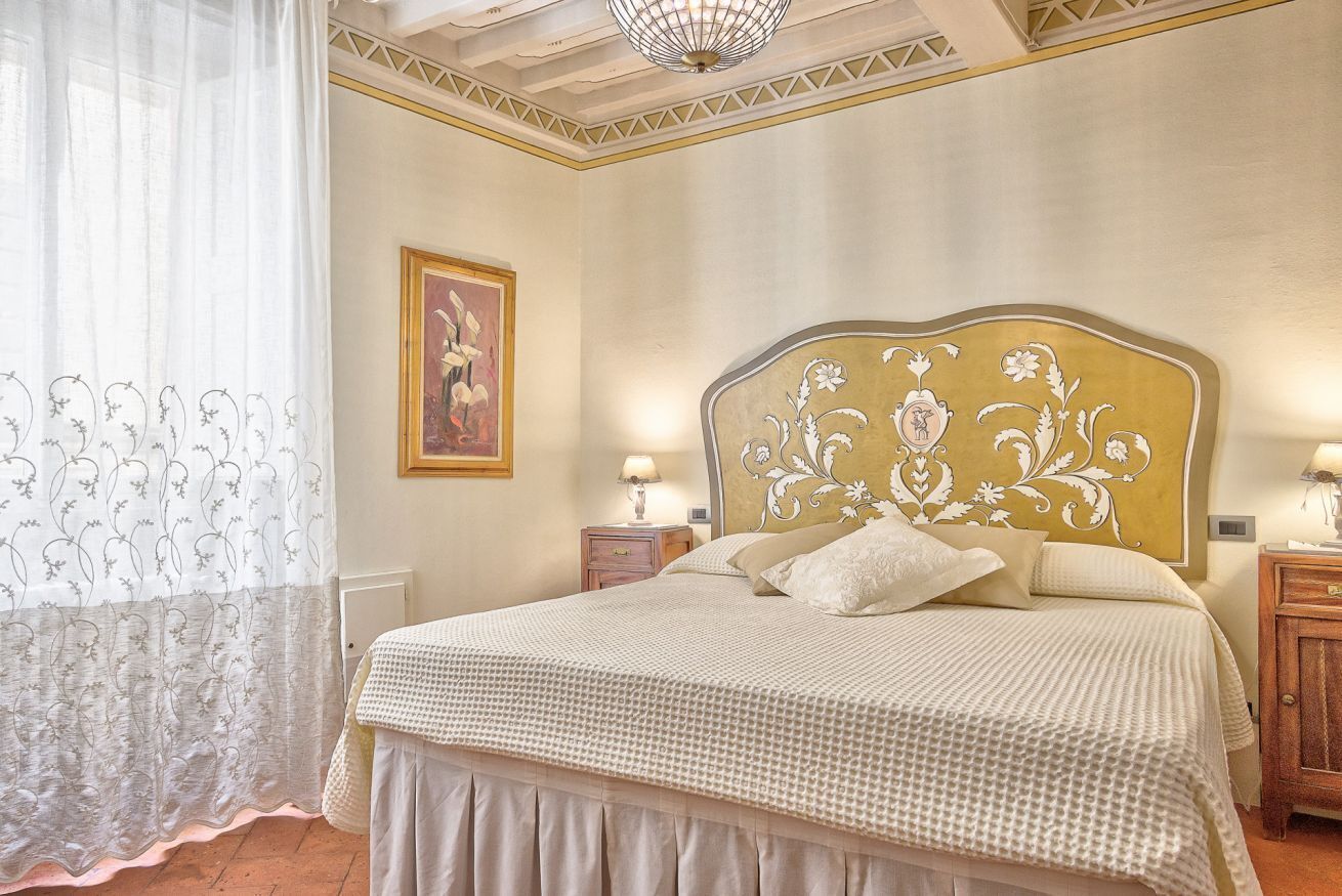 Una camera da letto con un letto grande, una testiera con motivo floreale, due comodini con lampade e una finestra con tende leggere.