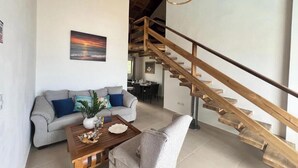 A modern living room with a wooden staircase, a grey sofa, a coffee table, and a painting of a sunset. - Las Terrenas Gorgeous Penthouse (Las Terrenas)