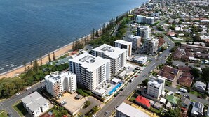An aerial view of a coastal residential area with modern buildings, a beach, and a clear blue sky. - Stylish beachside unit 3 min walk to beach (Redcliffe)