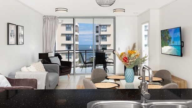 A modern living room with a sofa, chairs, a dining area, and a kitchen with a sink. - Stylish beachside unit 3 min walk to beach (Redcliffe)