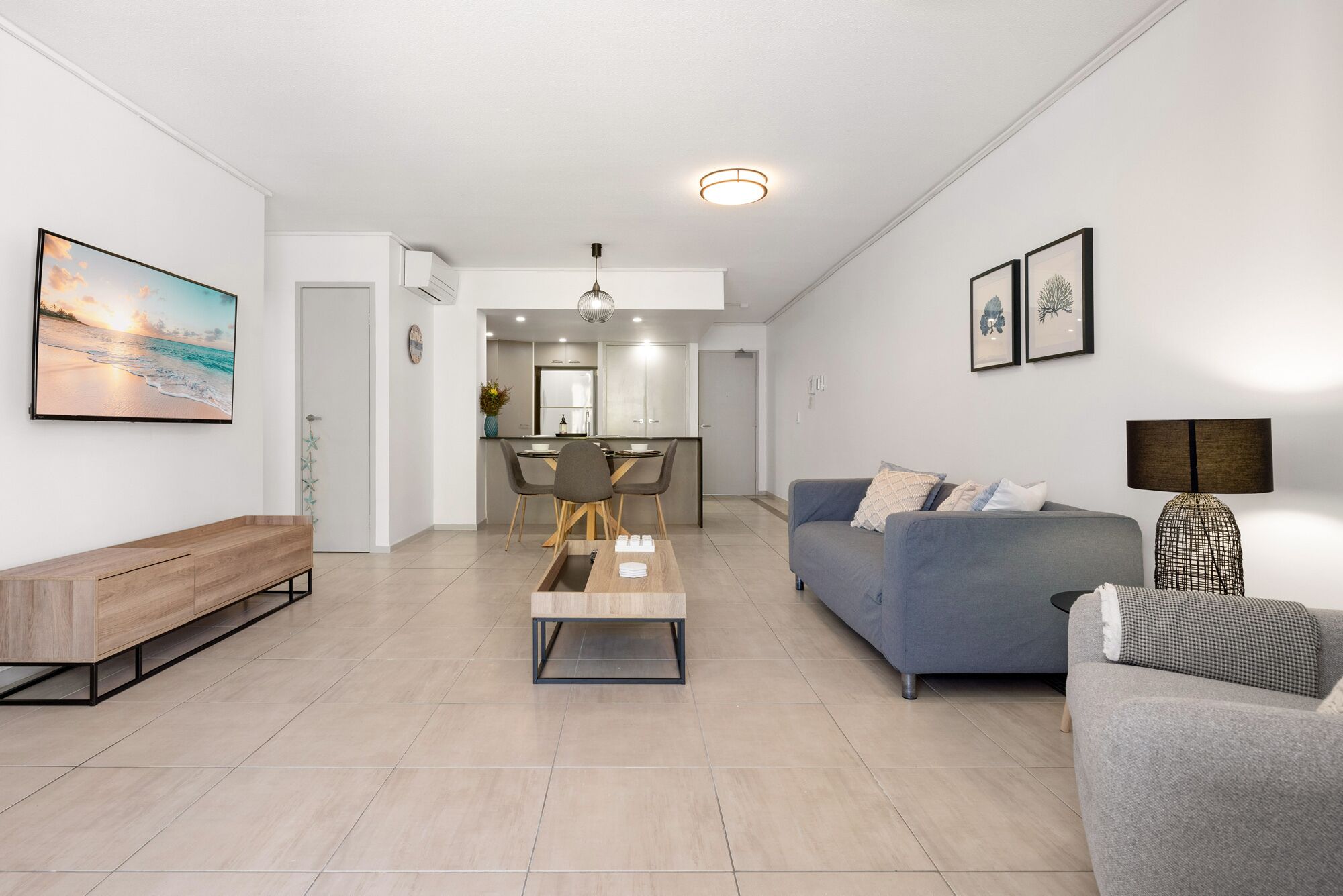 A modern living room with a sofa, a coffee table, and a bench. There is a dining area with a table and chairs. The room has white walls and a ceiling light. - Stylish beachside unit 3 min walk to beach (Redcliffe)
