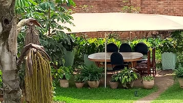 Un espace patio aménagé avec une table et des chaises sous une tonnelle, entouré de plantes en pot et d’un arbre.