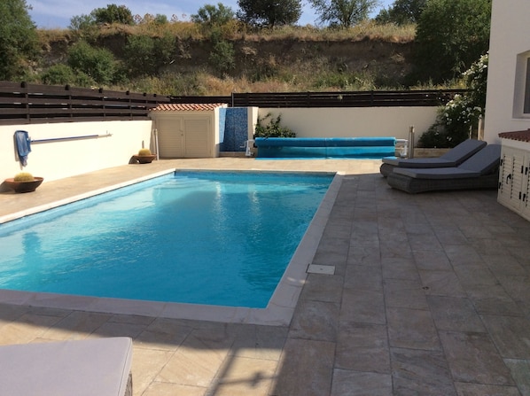 Une piscine rectangulaire bordée de carreaux, entourée d’une terrasse en pierre. On y trouve des chaises longues et une petite table sur laquelle est posée une plante succulente. Une clôture en bois et un bâtiment avec un toit recouvert de tuiles sont visibles à l’arrière-plan.
