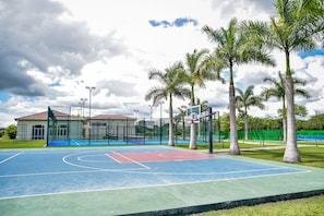 Un terrain de basket en plein air, doté d’une surface bleue et verte, entouré de palmiers et bordé d’un bâtiment avec une zone délimitée par une clôture.