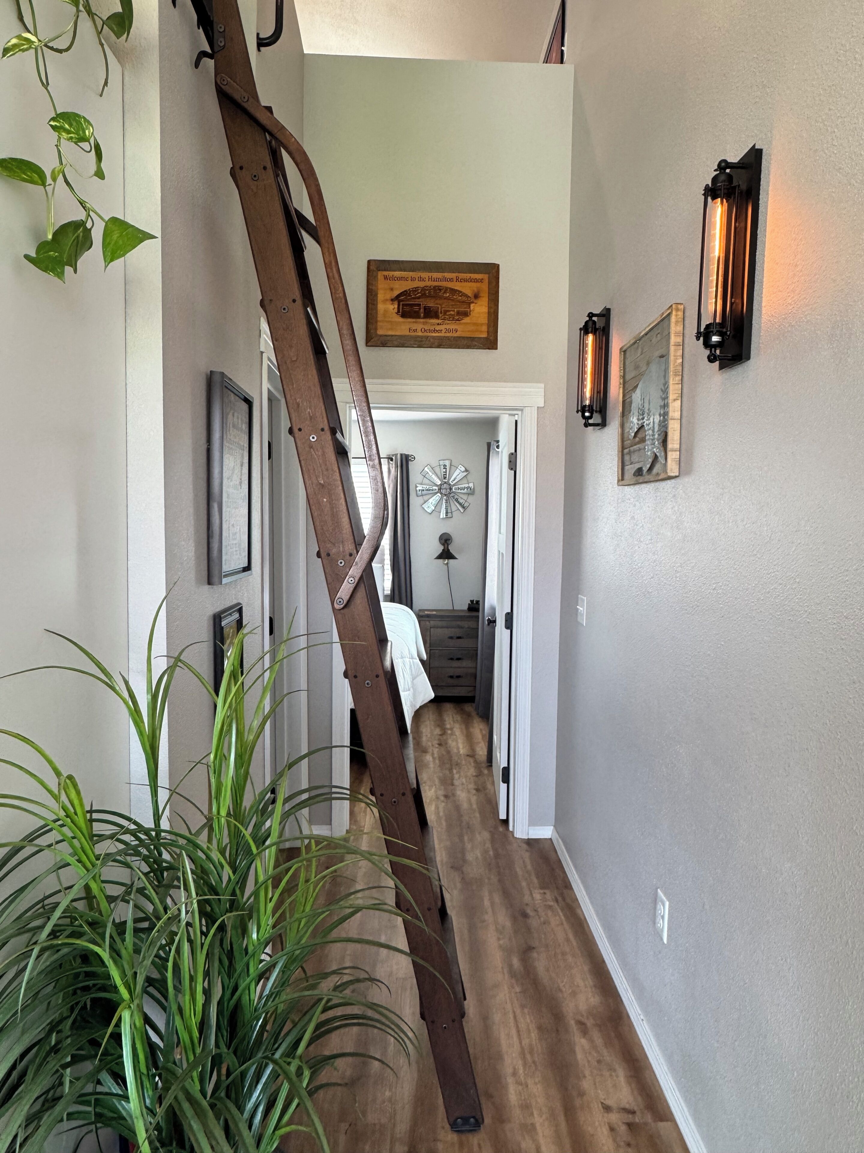 Un couloir avec une échelle en bois, une pancarte de bienvenue, des lampes fixées au mur et une porte donnant sur une chambre.