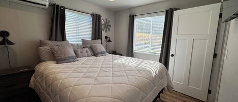 A bedroom with a large bed, two windows with blinds, a door, and an air conditioning unit.