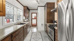 A kitchen with wooden cabinets, a stainless steel refrigerator, a stove, and a window with a view of a snowy landscape. - Third & Lake - Downtown Sandpoint (Sandpoint)