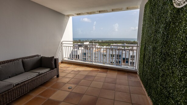 Ein Balkon mit einer Rattan-Couch, einer grünen Hecke und Blick auf Gebäude und Meer.