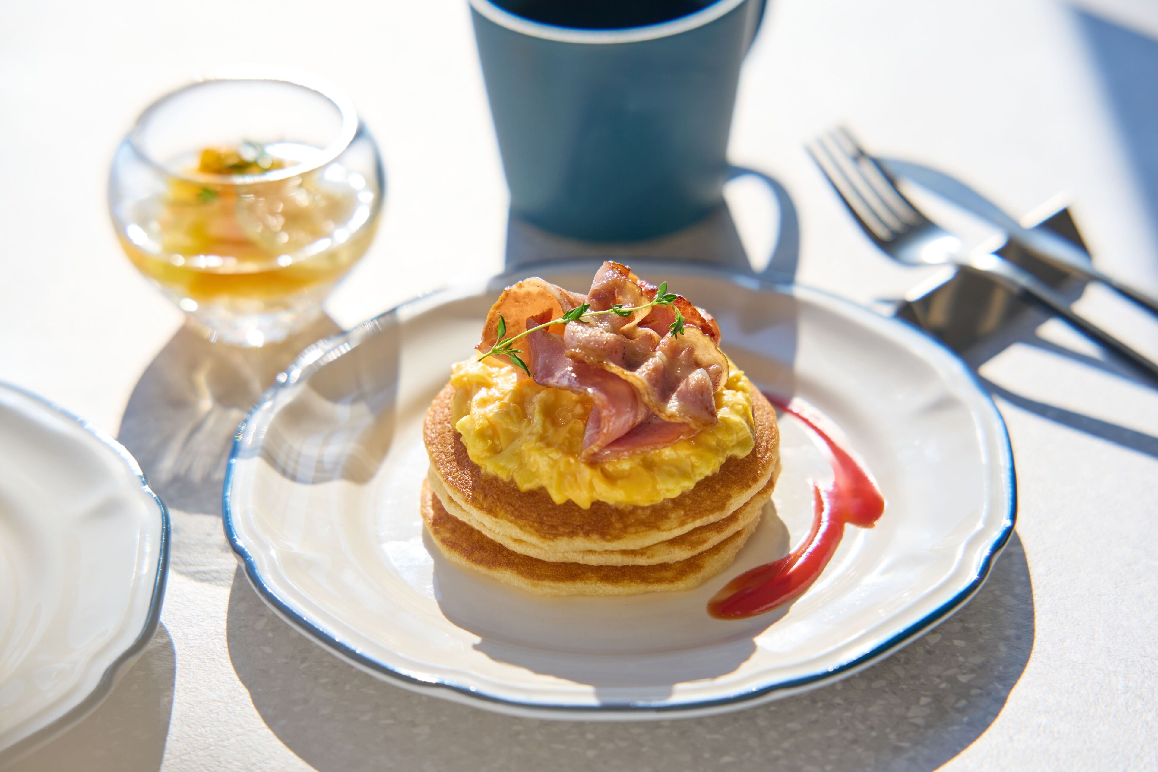 A breakfast plate with pancakes topped with ham and eggs, a small glass of a yellow beverage, and a blue cup in the background.
