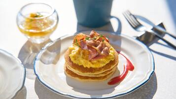 Un plato de desayuno con panqueques cubiertos de jamón y huevos, un vaso pequeño de una bebida amarilla y una taza azul al fondo.