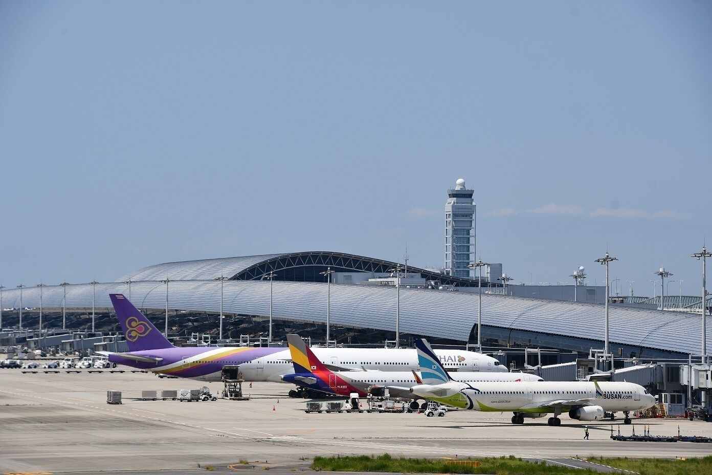 An airport with multiple airplanes parked, a large terminal building, and a control tower in the background.