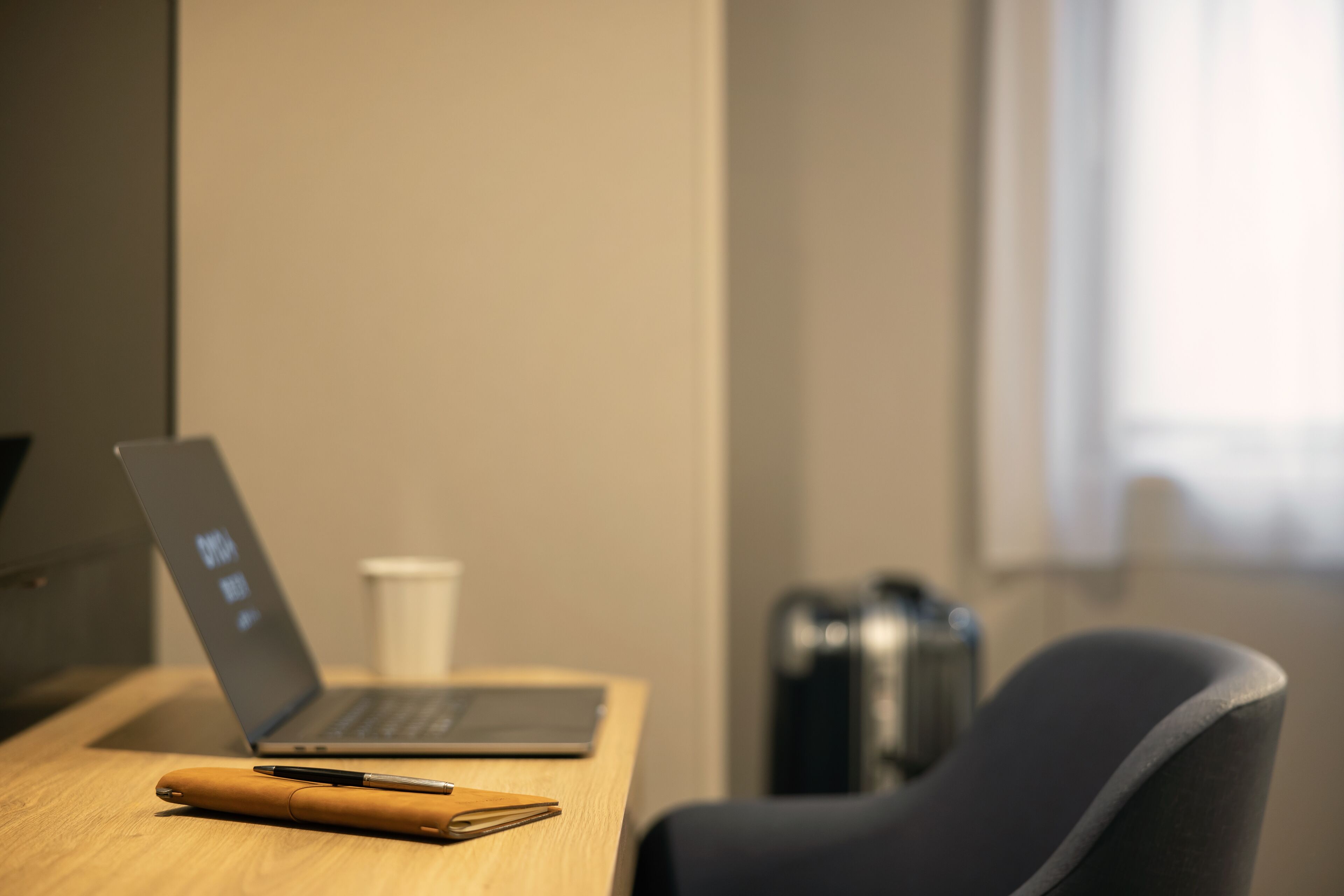 A laptop on a wooden desk with a pen and a notebook beside it, a white cup, and a black chair.