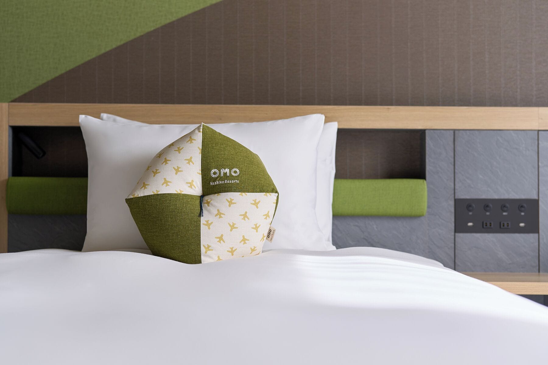 A hotel room with a bed, a headboard with a control panel, and a pillow with a geometric design.
