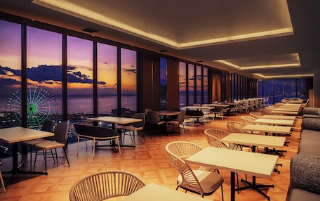 A modern restaurant with a view of the sunset over the sea, featuring high ceilings, large windows, and a variety of seating options.