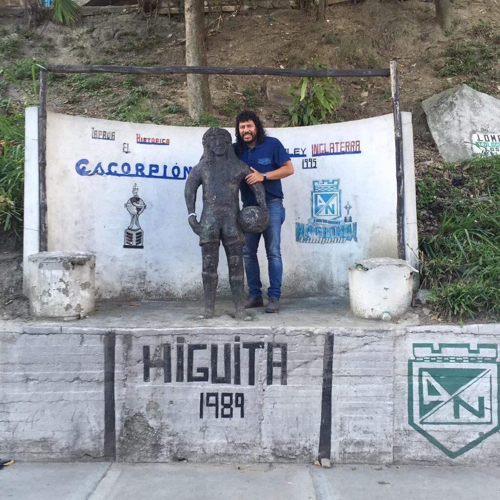 Un hombre está de pie junto a una estatua sobre un pedestal con un cartel que muestra el año 1989 y el nombre "HIGUITA".