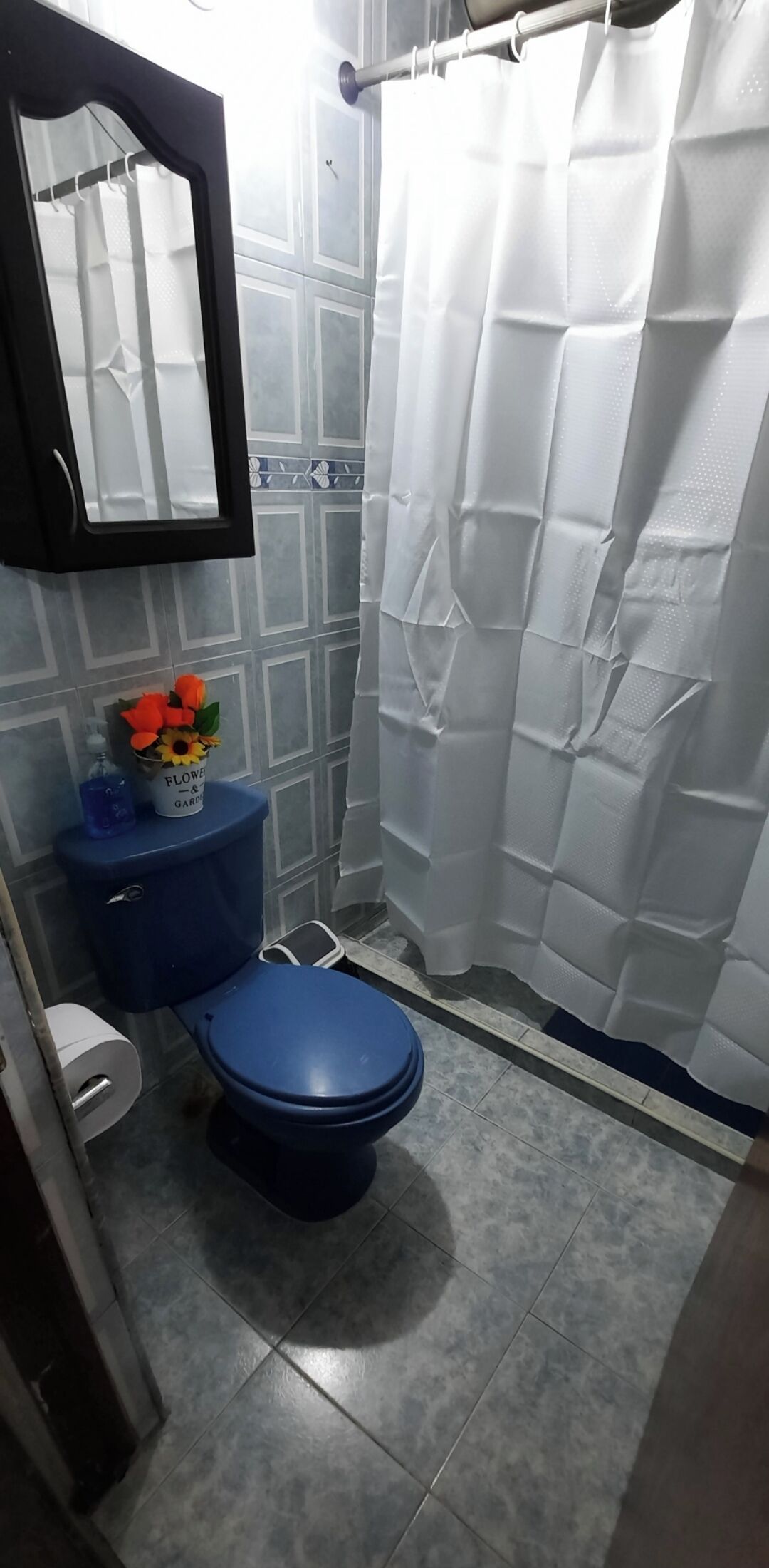 Un baño con inodoro azul, cortina de ducha blanca y un espejo sobre el lavabo.