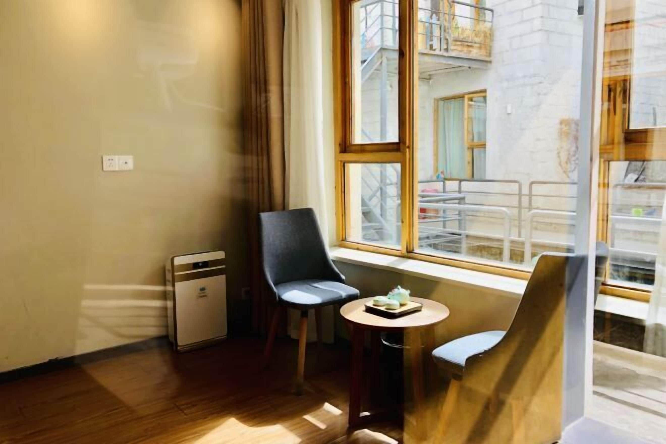 Una habitación con una pequeña mesa, dos sillas cerca de una ventana, un equipo de aire acondicionado compacto y un enchufe visible en la pared.