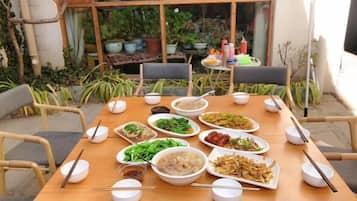 A dining table set with various dishes and bowls, surrounded by chairs, in an outdoor setting with plants and a building in the background.