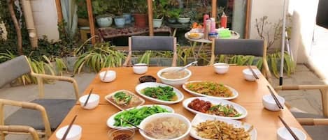 Mesa de comedor dispuesta con diferentes platos y tazones, rodeada de sillas, en un entorno exterior con plantas y un edificio al fondo.