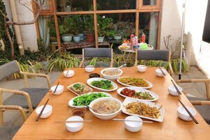 Mesa de comedor dispuesta con diferentes platos y tazones, rodeada de sillas, en un entorno exterior con plantas y un edificio al fondo.