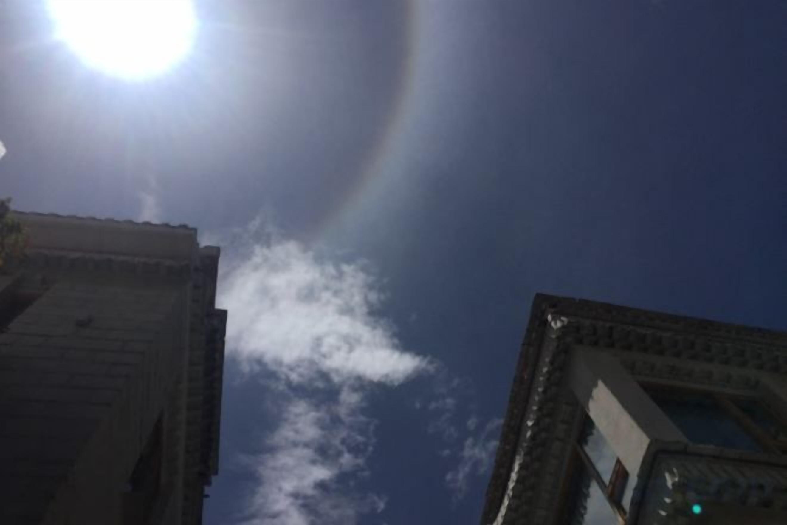 A clear sky with a sun visible through the clouds, and the tops of buildings.