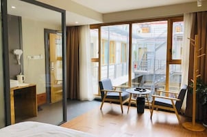 Una habitación de hotel con una cama, una mesita con sillas, un ventanal con vista a un edificio y un perchero.