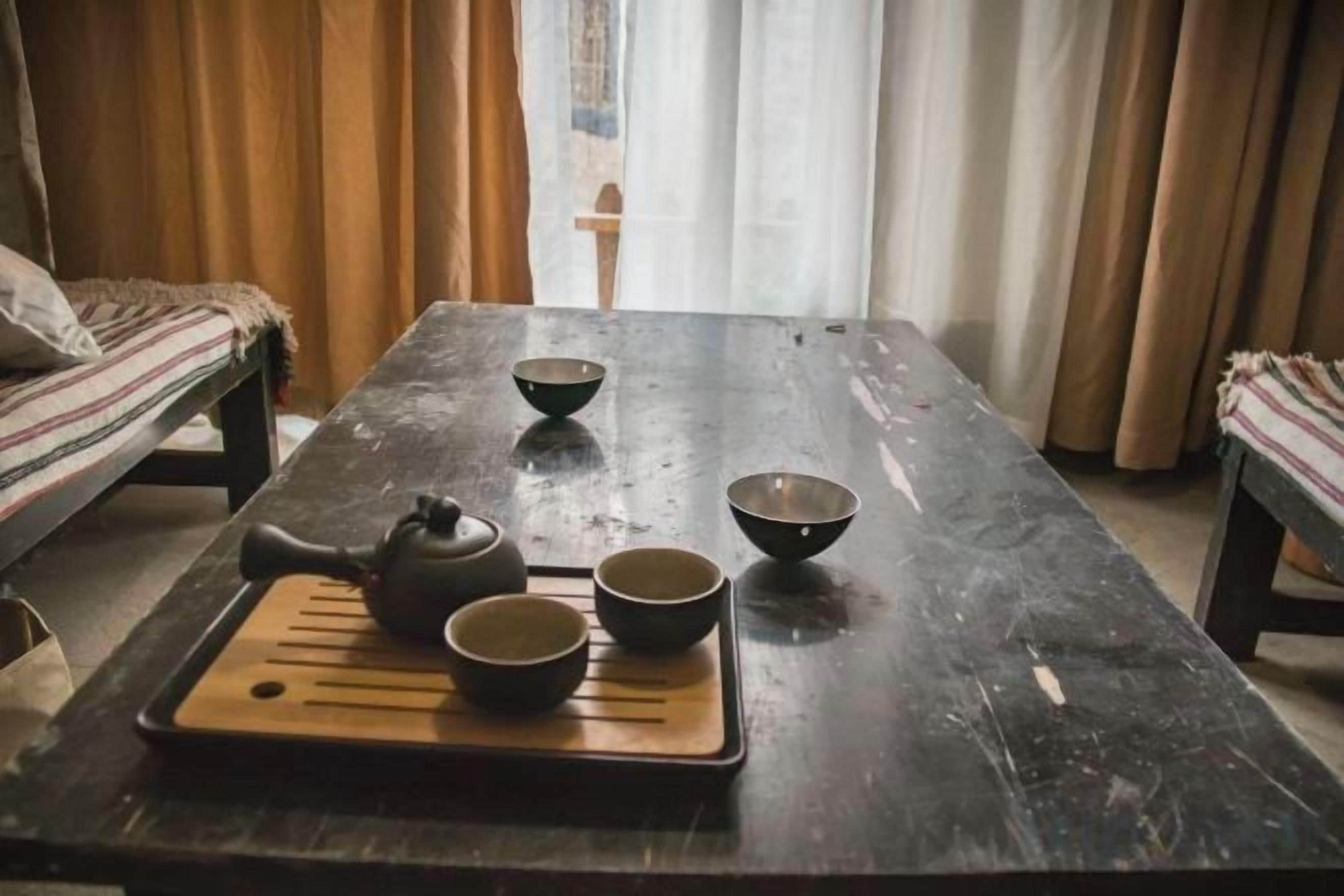 Una mesa de madera con una bandeja que sostiene una tetera y tazas, ubicada en una habitación con cortinas y un banco.