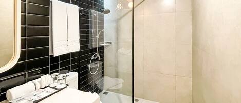 Un baño moderno con ducha envuelta en vidrio, un lavamanos blanco con artículos de aseo y una pared revestida de baldosas negras.