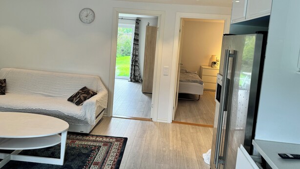 En moderne stue med en hvid sofa, et lille rundt bord og et mønstret tæppe. Der er et køkken med et køleskab og skabe. Et ur hænger på væggen.