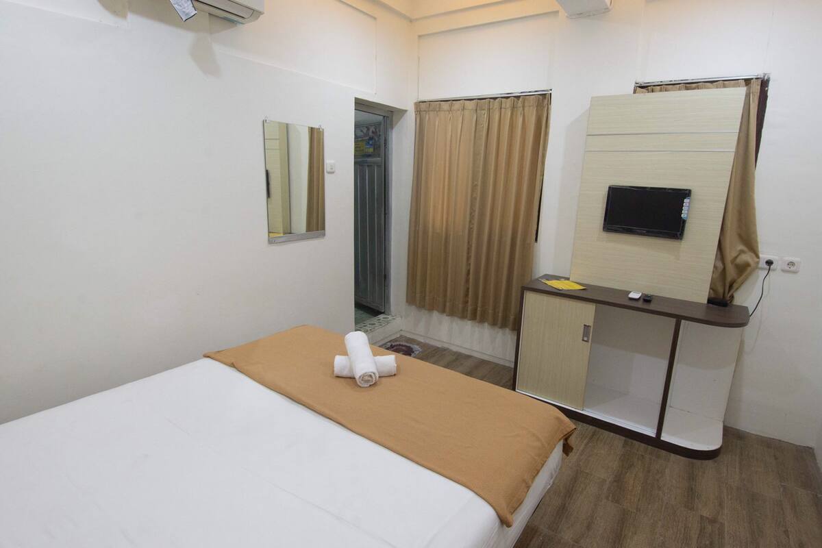 Quarto de hotel com cama, escrivaninha, espelho, televisão e zona de duche.