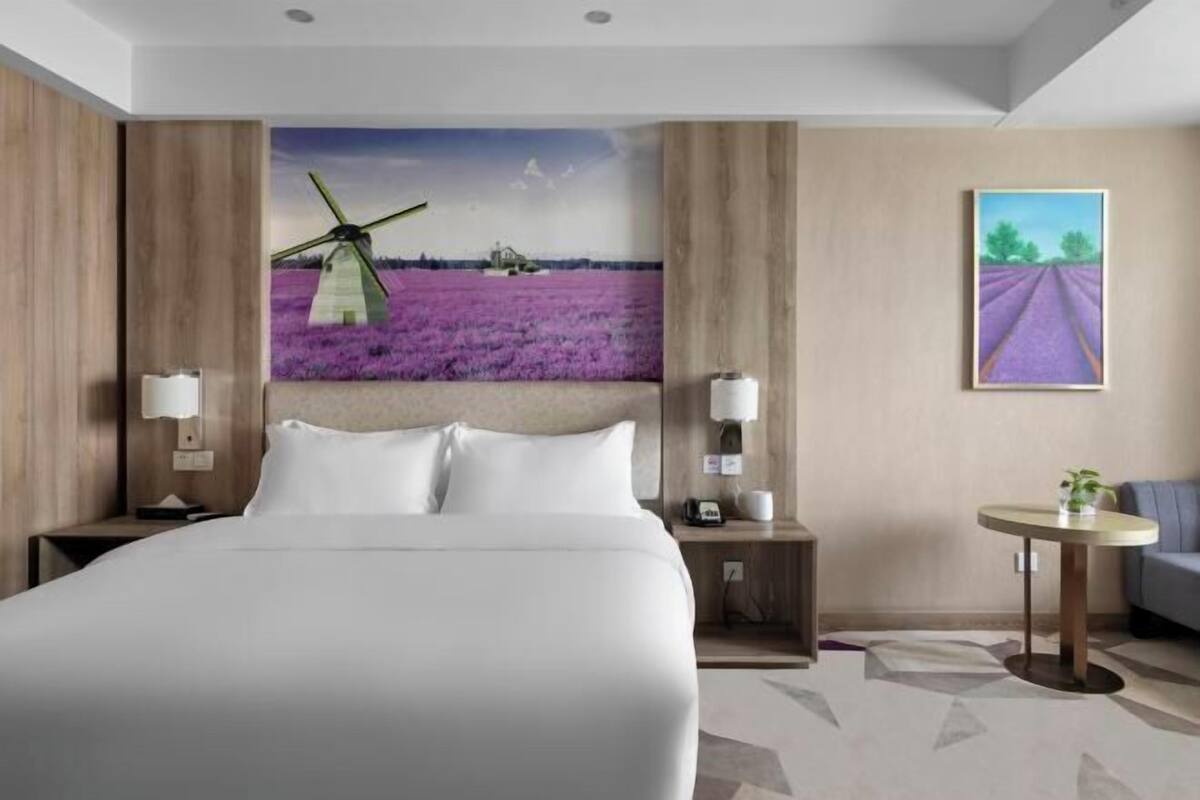 A hotel room with a large bed, a nightstand with a lamp, a small table with a plant, and a painting of a lavender field and windmill on the wall.