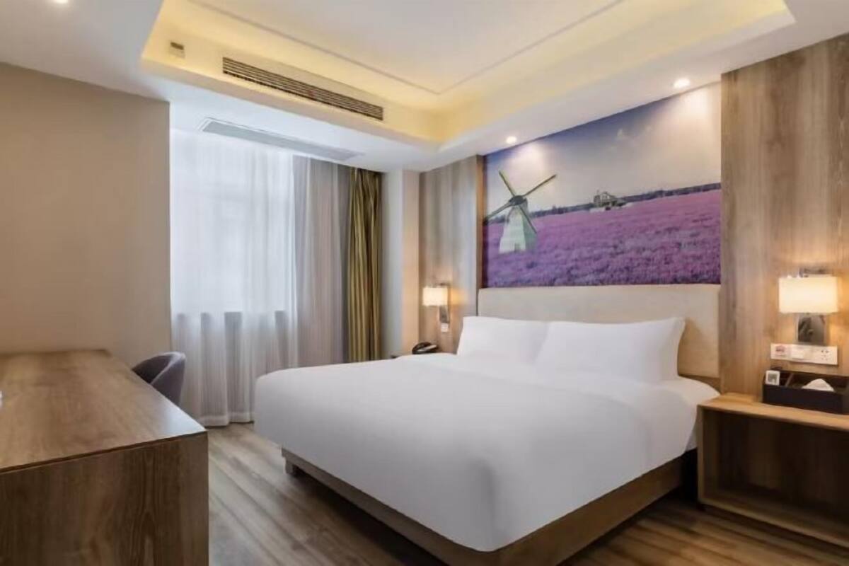 A hotel room with a large bed, a desk, a chair, a window with curtains, and a painting of a windmill on the wall.