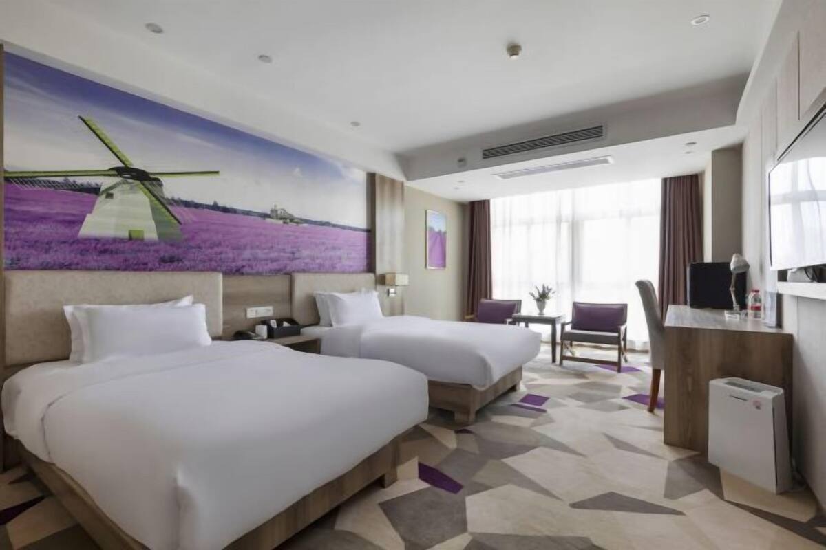 A hotel room with two beds, a desk, a chair, a TV, and a painting of a windmill.