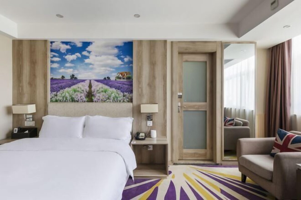 A hotel room with a bed, a painting of lavender fields, a sofa, and a door leading to another room.