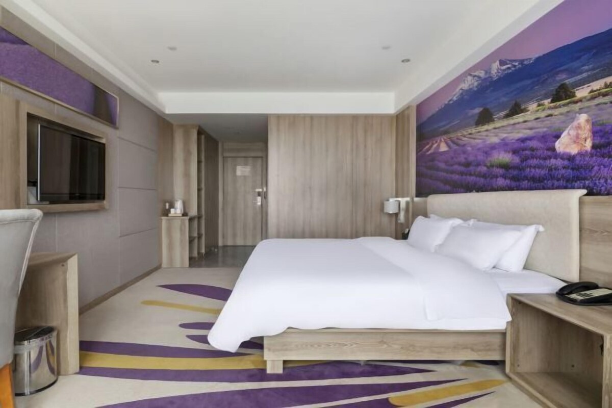 A hotel room with a large bed, a TV mounted on the wall, a wooden headboard, and a painting of a lavender field on the wall.