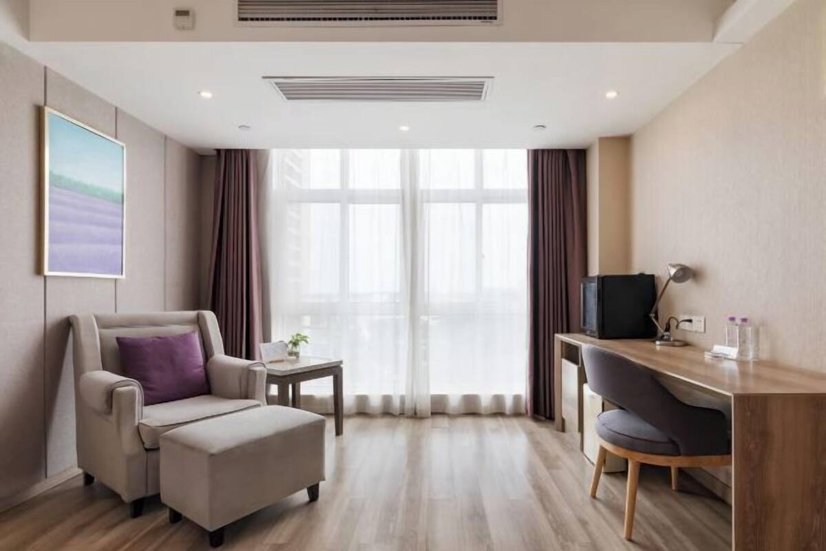 A modern hotel room with a sofa, a desk, a chair, a TV, and a large window with curtains.