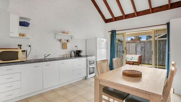 Une cuisine compacte avec des meubles blancs, un four à micro-ondes, une cuisinière et un réfrigérateur. On y trouve une table à manger avec des chaises, et une porte vitrée donne sur le jardin.