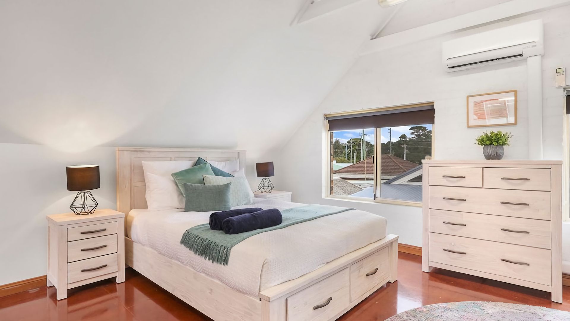 A bedroom with a bed, bedside tables, a dresser, a window with a view, and an air conditioning unit.