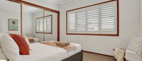 A bedroom with a bed, a chair, a large mirror, and a window with shutters.