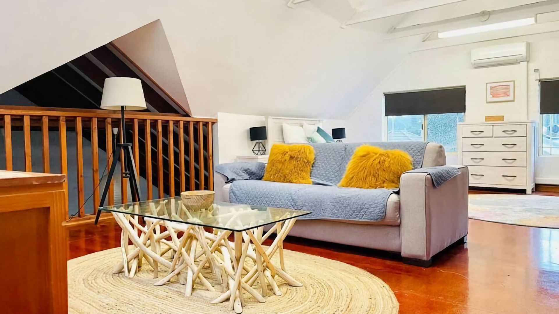 A modern living room with a glass coffee table, a sofa with yellow cushions, a wooden staircase, and a white dresser.
