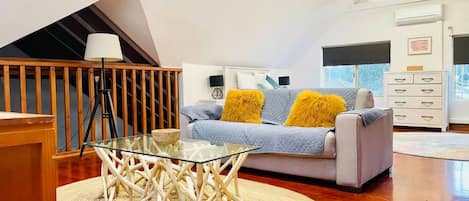 A modern living room with a glass coffee table, a sofa with yellow cushions, a wooden staircase, and a white dresser.