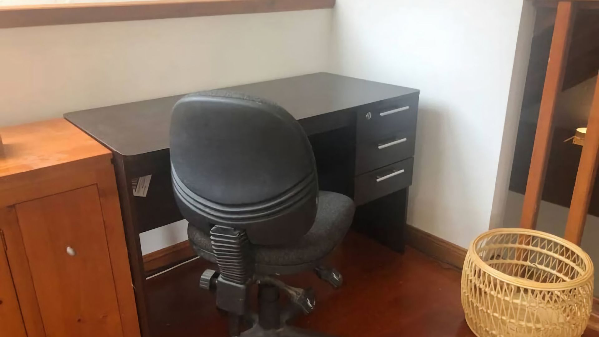 Une pièce comprenant un bureau en bois, un fauteuil de bureau noir, un panier en osier et une armoire en bois.