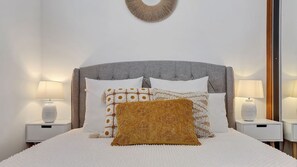 Un lit bien fait, doté d’une tête de lit rembourrée de couleur grise, de draps blancs et de plusieurs coussins aux motifs variés. Deux tables de chevet avec des lampes se trouvent de chaque côté du lit.