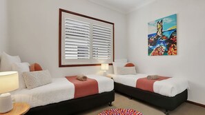 Ein Hotelzimmer mit zwei Betten, einem großen Fenster mit Jalousien, einem farbenfrohen Bild an der Wand und einem kleinen runden Teppich.